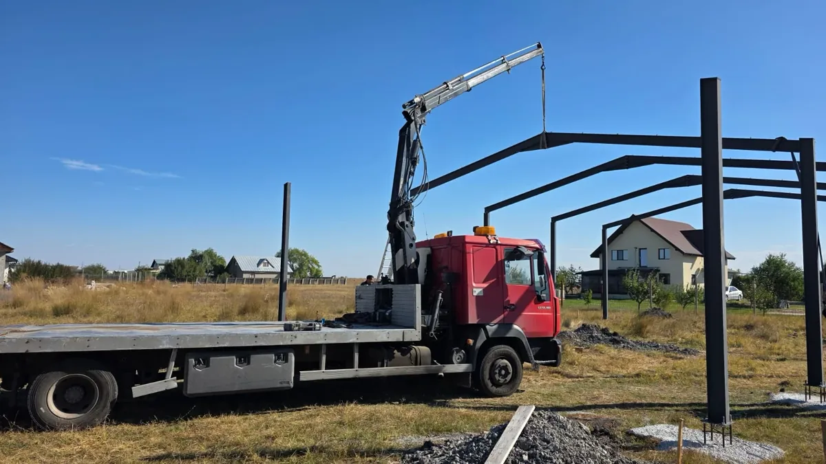 Camionul roșu cu macara ținând o grindă la montajul unei hale metalice