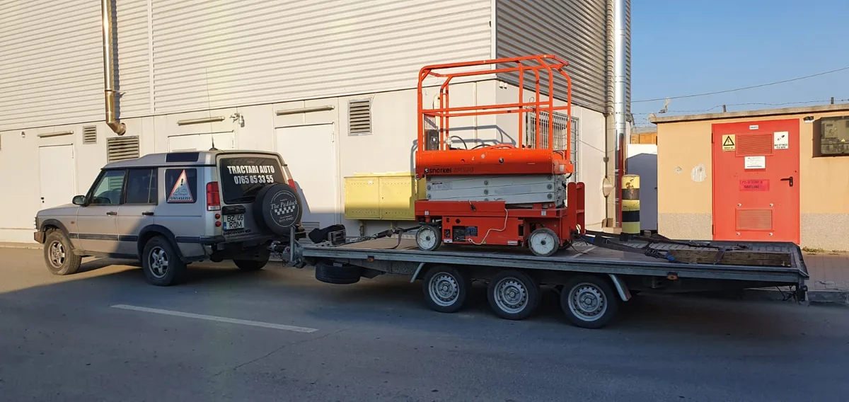 Nacelă foarfecă portocalie pe remorca-platformă, lângă o hală industrială